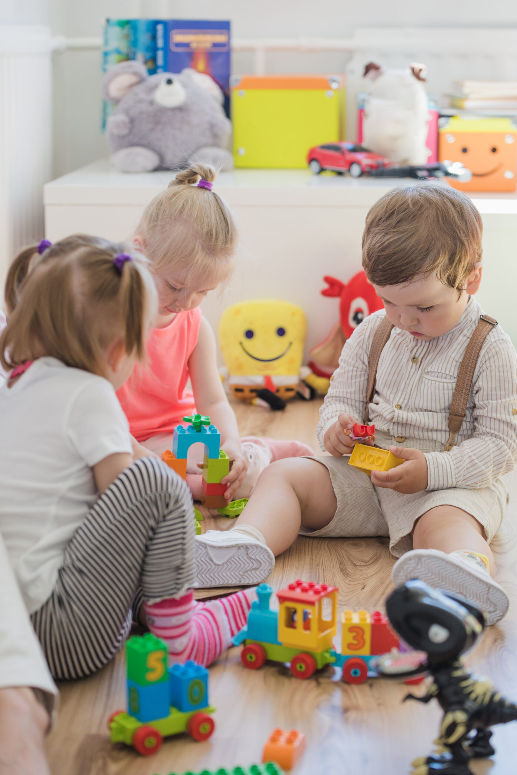 Dzieci bawiące się zabawkami, chłopiec trzyma w ręku dwa klocki, dwie dziewczynki bawią się w układanie zamku z klocków.